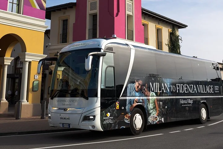 A silver and black Milan to Fidenza Village coach parked outside pink and yellow buildings